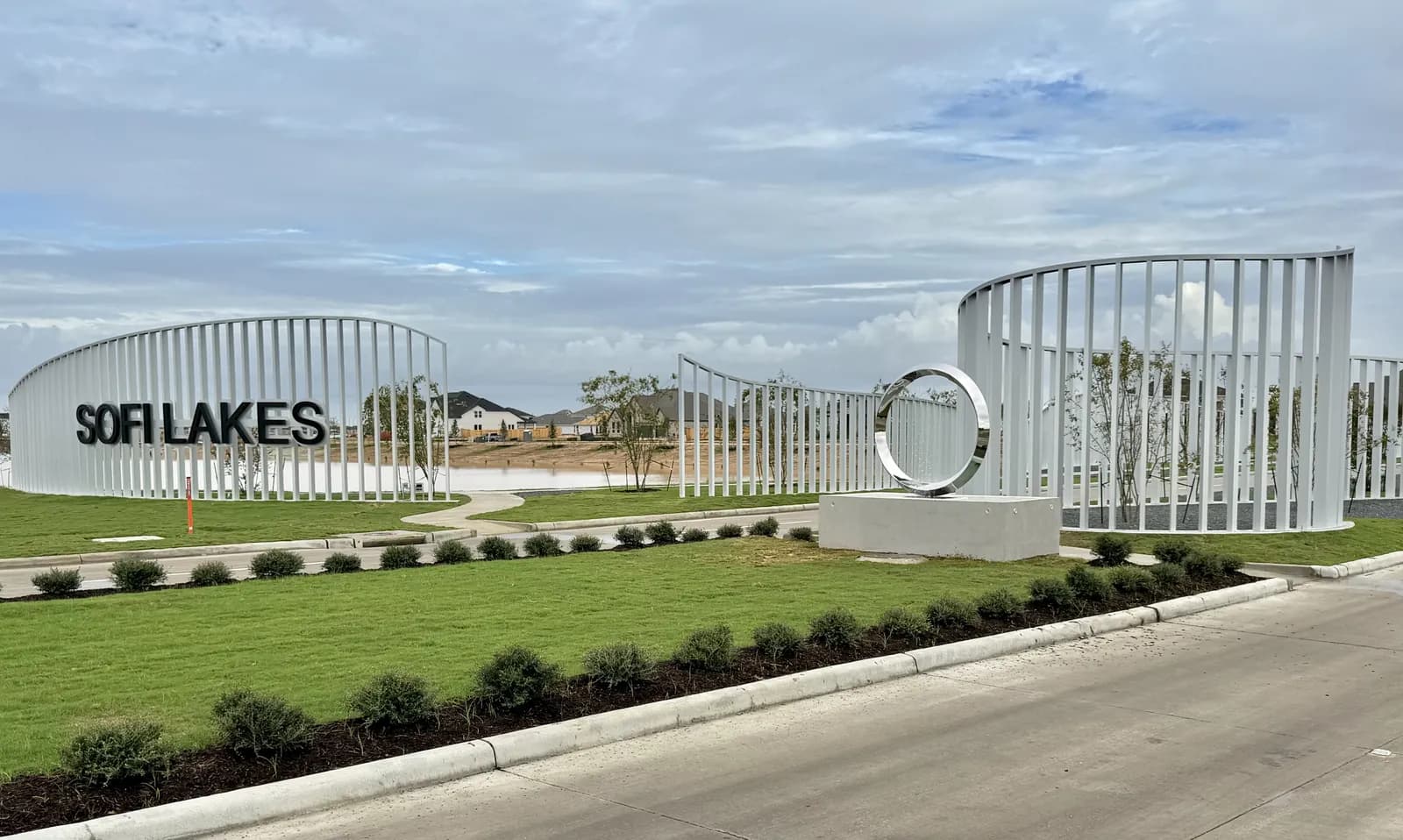 Sofi Lakes main entry monument with curved white slat walls, Sofi Lakes lettering and a stainless steel ring sculpture along the landscaped entry drive in Katy, TX